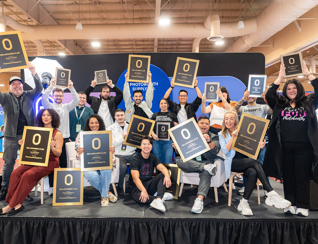 Team members of Photobooth Supply Co. smiling and holding up framed "Millionaires Club" and "Six Figure Club" awards. 